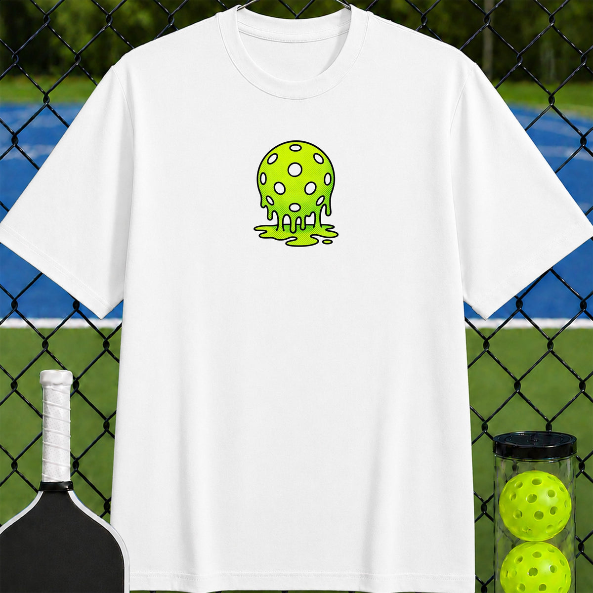 A white short-sleeve crewneck t-shirt featuring a green dripping slime skull graphic on the chest, displayed against a blurred background of a chain-link fence and tennis court with a pickleball racket and balls.